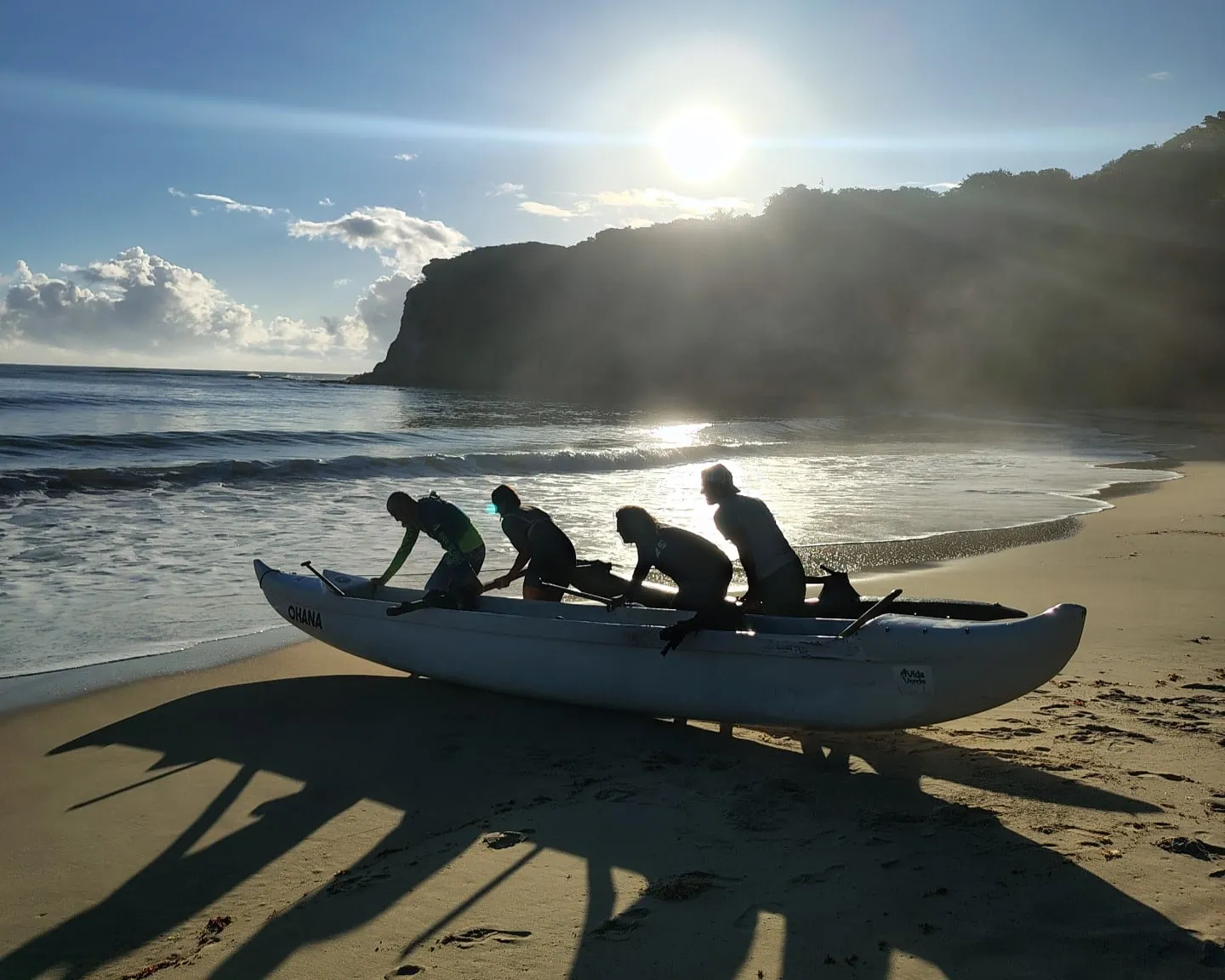Quatro remadores empurram uma canoa havaiana da areia para o mar em Pipa, com falésia e sol ao fundo