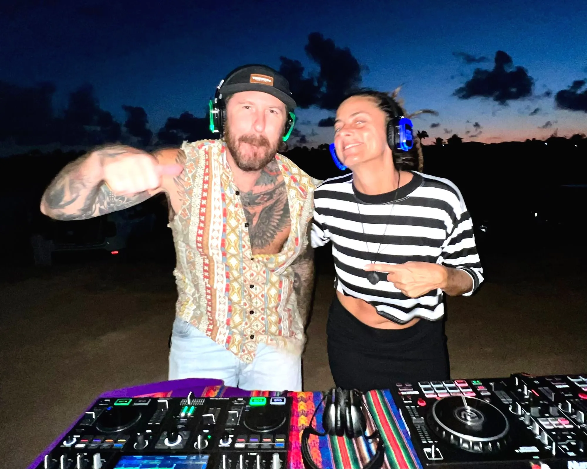 Dupla de DJs sorrindo ao anoitecer, com fones acesos em verde e azul, em Pipa
