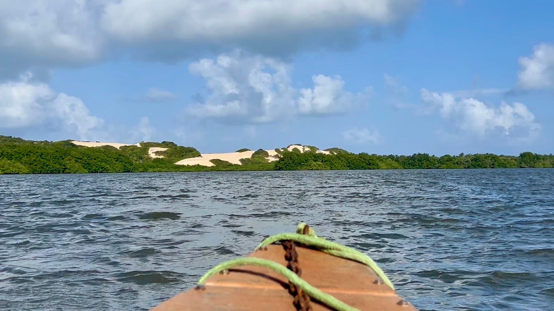 Proa de barco navegando na Lagoa Guaraíras com dunas ao fundo