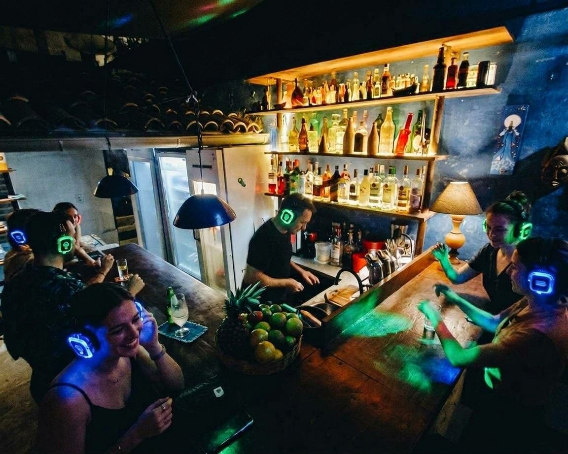 Pessoas com fones de silent disco acesos em azul e verde no balcão de drinks da Fonte, na Praia da Pipa